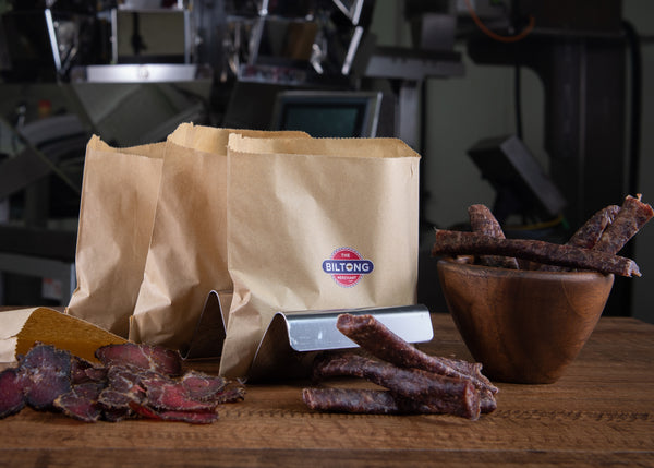fresh cut biltong and dröewors on a table with a wooden bowl and The Biltong Merchant branded paper bags