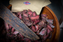 The Biltong Merchant logo on paper snack packs of fresh cut lean biltong with a bowl of droëwors