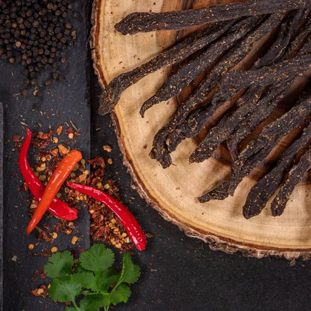 Traditional meat beer sticks stokkies on a wooden board with, spices, peri peri chilli flakes & chillies on slate background