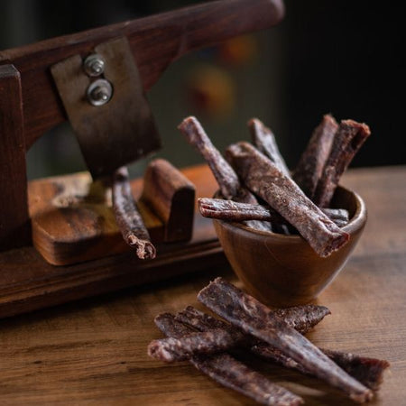 Tasty traditional droëwors sausages in a small wooden bowl on a table with vintage wooden biltong cutter in the background