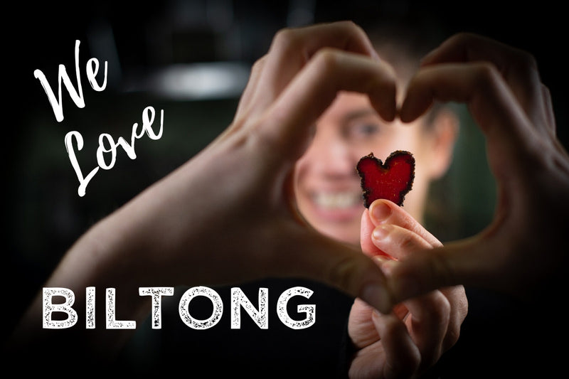 Smiling woman holds heart-shaped slice of biltong with two hands creating a heart: The Biltong Merchant - We Love Biltong!